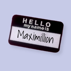 Puede incluir: Una etiqueta con el nombre en blanco y negro con el texto "HELLO my name is Maximillion". La etiqueta tiene forma rectangular con esquinas redondeadas y un centro blanco.
