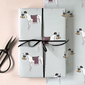 May include: A rectangular gift wrapped in light blue paper featuring a repeating pattern of white greyhounds wearing striped blankets and carrying gold bells in their mouths, tied with a thin black ribbon, accompanied by black metal scissors.