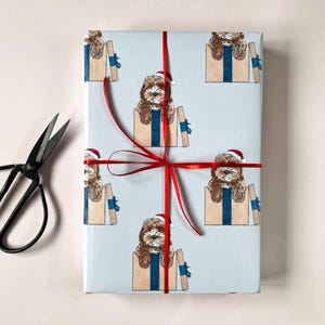 May include: A rectangular gift box wrapped in light blue paper featuring a repeating illustration of a brown and white dog wearing a red Santa hat and sitting inside a tan gift box, tied with a thin red ribbon and accompanied by black scissors.
