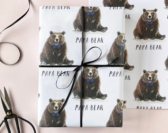 May include: Gift-wrapped present featuring wrapping paper with a repeating pattern of a brown bear wearing a blue bow tie, with the text "PAPA BEAR". The gift is tied with a black ribbon, with scissors and a ribbon roll in the background.