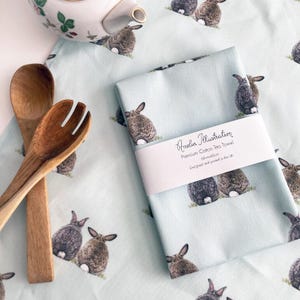 Könnte beinhalten: Hellblaues Baumwoll-Geschirrtuch mit einem sich wiederholenden Muster aus braunen und grauen Hasen. Das Handtuch ist mit einem weißen Band umwickelt, auf dem steht "Amelia Illustration Premium Cotton Tea Towel 58cm x 45cm Designed and printed in the UK". Holzlöffel und -gabel im Bild.
