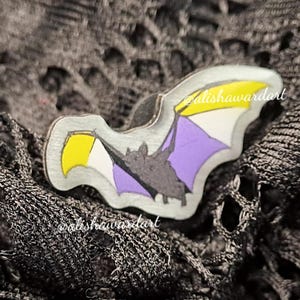 May include: A silver bat-shaped pin featuring the colours of the non-binary pride flag. The wings are purple, white, and yellow. The pin is set against a dark, textured background. The artist's name, alishawardart, is visible.