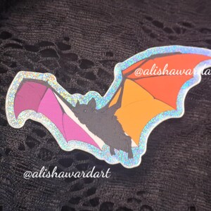 May include: A sticker of a bat with wings in shades of orange, pink, and purple. The bat has a dark grey body and is outlined with a shimmering, iridescent border. The text "@alishawart" is visible on the sticker.