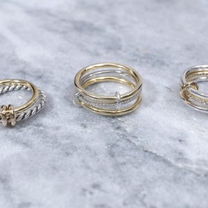 May include: Three gold and silver rings. The ring on the left is a twisted band of silver with a gold accent. The middle ring is a stack of three bands, two silver and one gold. The ring on the right is a silver band with a gold accent and a row of small diamonds.