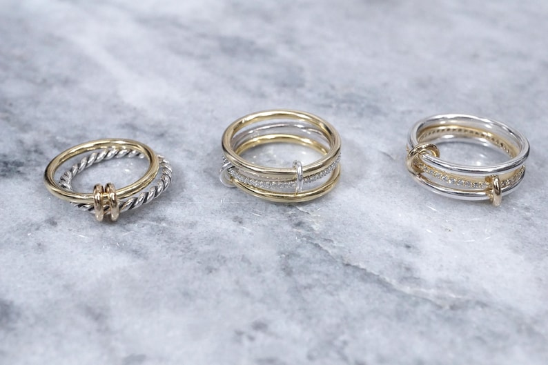 May include: Three gold and silver rings. The ring on the left is a twisted band of silver with a gold accent. The middle ring is a stack of three bands, two silver and one gold. The ring on the right is a silver band with a gold accent and a row of small diamonds.