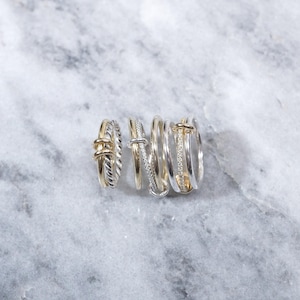 May include: A set of six silver and gold rings stacked together. The rings are all different styles, with some featuring a twisted design and others with a simple band. The rings are on a white marble background.