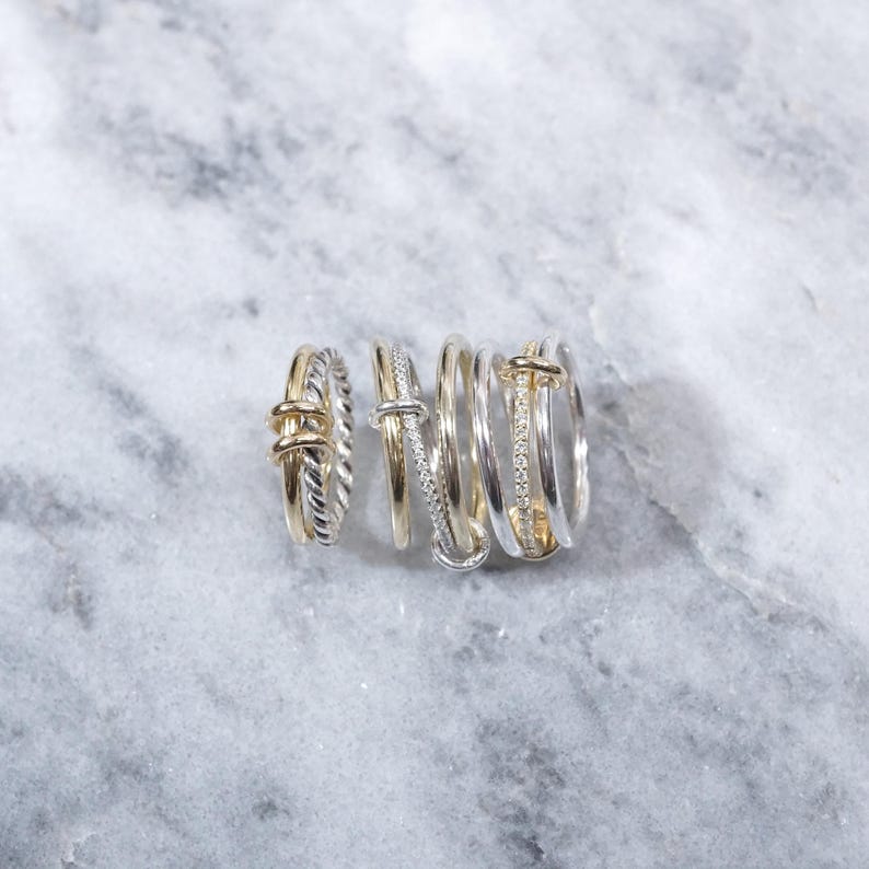 May include: A set of six silver and gold rings stacked together. The rings are all different styles, with some featuring a twisted design and others with a simple band. The rings are on a white marble background.