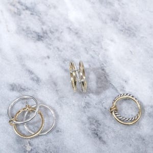 May include: Three gold and silver rings on a marble surface. One ring is a double band with a small diamond, another is a single band with a twisted design, and the third is a set of three interlocking bands.