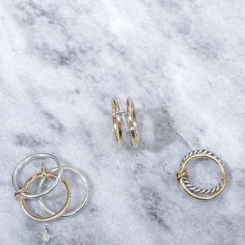 May include: Three gold and silver rings on a marble surface. One ring is a double band with a small diamond, another is a single band with a twisted design, and the third is a set of three interlocking bands.