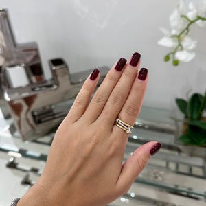 May include: A hand with dark red nail polish is wearing a gold and silver ring with small diamonds. The ring is on the middle finger.