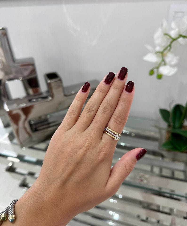 May include: A hand with dark red nail polish is wearing a gold and silver ring with small diamonds. The ring is on the middle finger.
