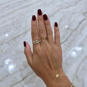 May include: A woman's hand with dark red nail polish is wearing a gold chain bracelet with a heart charm and three gold beaded bracelets. The hand is also wearing three gold rings on the ring finger, including a thin band with diamonds and a thicker band with a design.