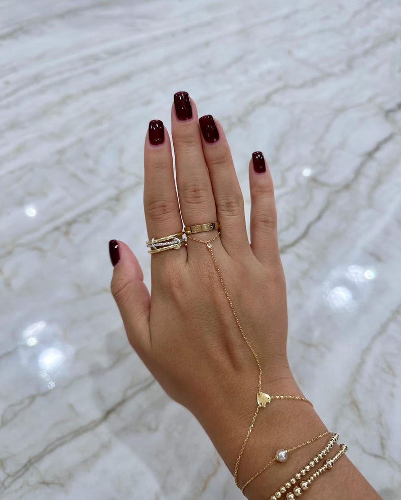 May include: A woman's hand with dark red nail polish is wearing a gold chain bracelet with a heart charm and three gold beaded bracelets. The hand is also wearing three gold rings on the ring finger, including a thin band with diamonds and a thicker band with a design.