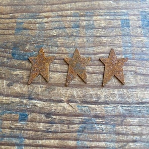 May include: Three rustic, star-shaped metal ornaments with a weathered, rusty finish. The stars are arranged horizontally against a wooden background with visible grain and faded blue text.