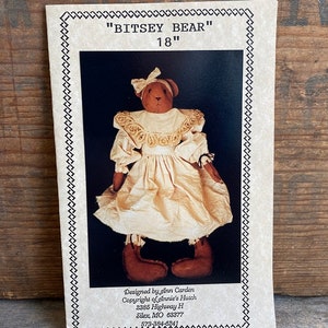 May include: A paper pattern for an 18" Bitsey Bear doll. The cover features a brown fabric bear wearing a cream-colored dress with a ruffled collar and a matching bow on its head. Text includes "BITSEY BEAR", "18"", and designer/copyright information.