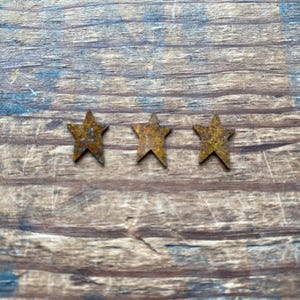 May include: Three small, rusty metal stars arranged horizontally against a weathered, brown wooden background. The stars have a textured appearance, suggesting an aged or vintage aesthetic. The wood grain adds a rustic element.