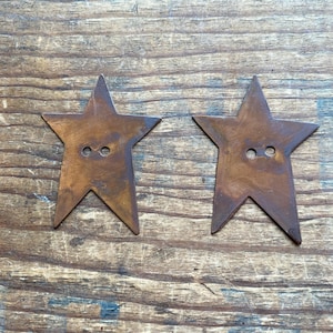 May include: Two rustic, star-shaped buttons with a distressed, bronze finish. Each button has two small holes for sewing. The buttons are displayed on a weathered wooden surface, suggesting a vintage or handcrafted aesthetic.
