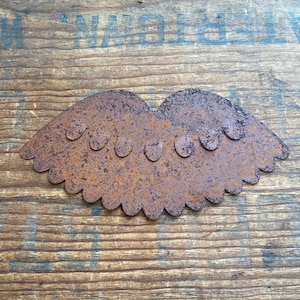 May include: A rusty brown metal decorative piece in the shape of a stylized pair of lips with a scalloped edge. The lips have six small, rounded indentations across the top. The piece is set against a wooden background.