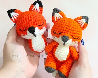 May include: Two crocheted fox plushies, one orange and white with closed eyes and the other orange and white with closed eyes. The plushies are held in the hands of a person.