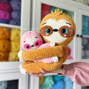Peut inclure: Un grand jouet en peluche de paresseux crocheté tenant un plus petit jouet en peluche de paresseux crocheté. Le grand paresseux est brun avec des accents blancs et des yeux verts. Le petit paresseux est rose avec des accents blancs et des yeux roses. Le texte "@allfromjade" est visible en arrière-plan.