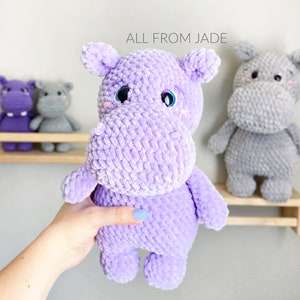 May include: A purple crocheted hippopotamus plush toy with blue eyes. The hippo is being held in a hand. The background shows two smaller hippopotamus plush toys on a shelf.