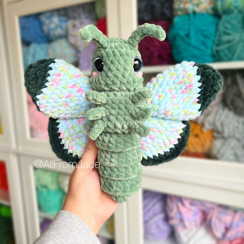 K&ouml;nnte beinhalten: Ein gr&uuml;nes, geh&auml;keltes Schmetterlings-Pl&uuml;schtier mit blauen und wei&szlig;en, floral gemusterten Fl&uuml;geln. Das Spielzeug hat gro&szlig;e schwarze Augen und eine kleine, geh&auml;kelte Antenne.