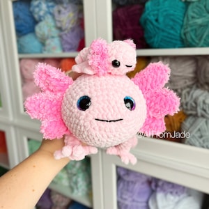 Peut inclure: Un jouet en peluche axolotl en crochet rose avec un plus petit jouet en peluche axolotl rose sur la tête. Le plus grand axolotl a des yeux bleus et un corps rose et duveteux. Le plus petit axolotl a des yeux noirs et un corps rose et duveteux. Le texte "@AllFromJade" est visible dans le coin inférieur droit de l'image.