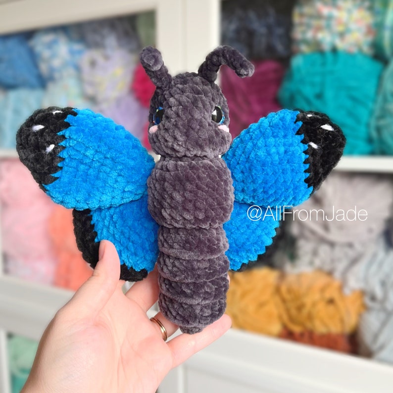 K&ouml;nnte beinhalten: Ein geh&auml;keltes Schmetterlings-Pl&uuml;schtier mit grauem K&ouml;rper und blauen Fl&uuml;geln. Das Spielzeug hat schwarze Akzente an den Fl&uuml;geln und ein kleines, rosafarbenes Gesicht.