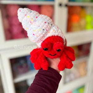 May include: A crocheted red hermit crab with a white and pink speckled shell. The crab is holding onto a hand. The background is a blurry image of a shelf with yarn.