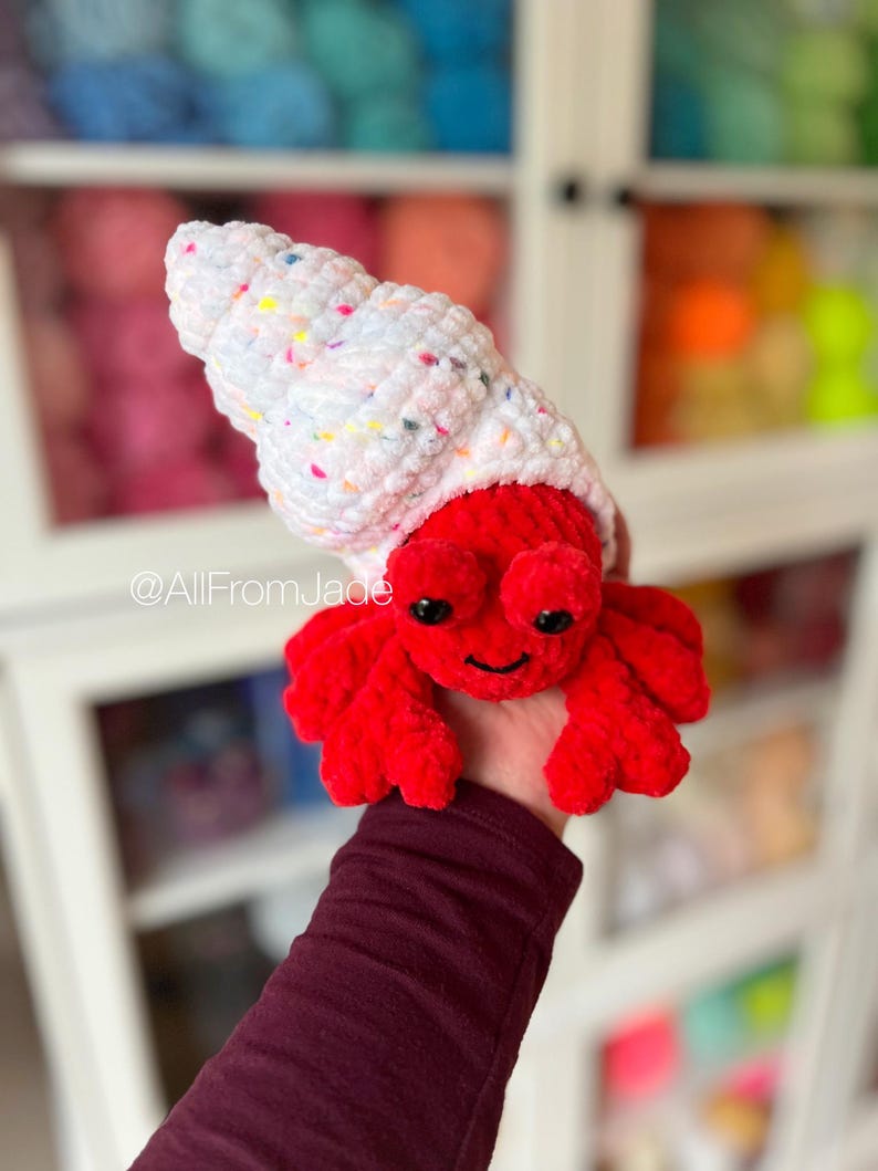 May include: A crocheted red hermit crab with a white and pink speckled shell. The crab is holding onto a hand. The background is a blurry image of a shelf with yarn.