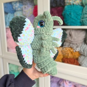 K&ouml;nnte beinhalten: Ein gr&uuml;nes, geh&auml;keltes Schmetterlings-Pl&uuml;schtier mit blauen Augen und rosa Wangen. Die Fl&uuml;gel sind mit einem Blumenmuster verziert.