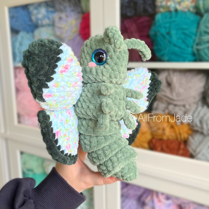 K&ouml;nnte beinhalten: Ein gr&uuml;nes, geh&auml;keltes Schmetterlings-Pl&uuml;schtier mit blauen Augen und rosa Wangen. Die Fl&uuml;gel sind mit einem Blumenmuster verziert.
