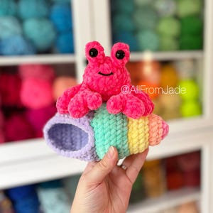 May include: A pink crocheted hermit crab toy with black eyes sitting in a rainbow striped crocheted shell. The shell is purple, green, yellow, and orange.  @AllFromJade