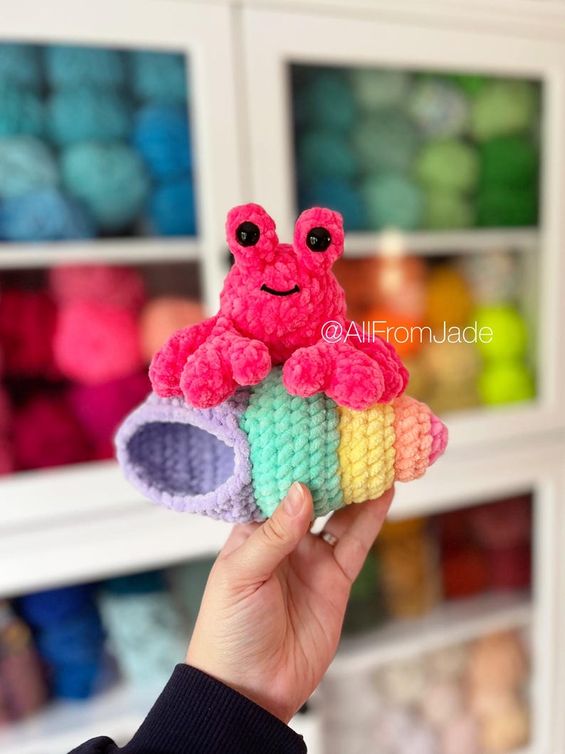May include: A pink crocheted hermit crab toy with black eyes sitting in a rainbow striped crocheted shell. The shell is purple, green, yellow, and orange.  @AllFromJade
