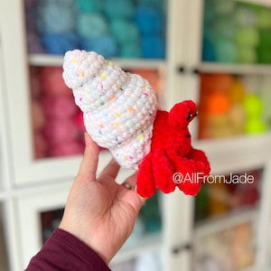 May include: A crocheted hermit crab toy with a white shell with colorful sprinkles and a red body. The toy is being held in a hand against a background of yarn.