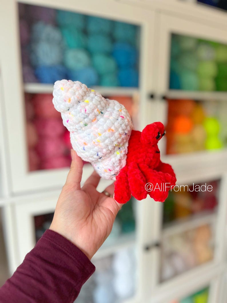 May include: A crocheted hermit crab toy with a white shell with colorful sprinkles and a red body. The toy is being held in a hand against a background of yarn.