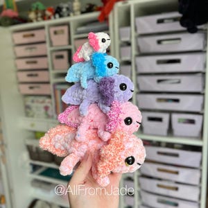 Op de afbeelding: Een stapel van zes handgemaakte gehaakte axolotls in verschillende pastelkleuren, waaronder roze, oranje, blauw en paars. Elke axolotl heeft zwarte veiligheidsoogjes en een franjerig, getextureerd ontwerp. De axolotls zijn op elkaar gestapeld.