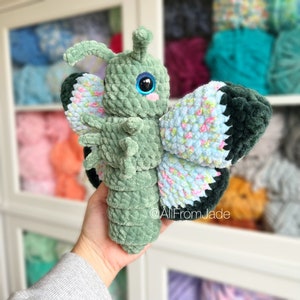 K&ouml;nnte beinhalten: Ein gr&uuml;nes, geh&auml;keltes Schmetterlings-Pl&uuml;schtier mit blauen und rosa floralen Fl&uuml;geln. Der Schmetterling hat gro&szlig;e, blaue Augen und eine rosa Nase.