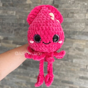 K&ouml;nnte beinhalten: Ein handgefertigtes, pl&uuml;schiges, pinkfarbenes H&auml;kelsquid-Spielzeug mit gro&szlig;en, glitzernden Augen und einem l&auml;chelnden Gesicht. Der Tintenfisch hat einen abgerundeten Kopf, kleine wei&szlig;e Akzente und lange, h&auml;ngende Tentakel. Das Spielzeug wird gehalten.