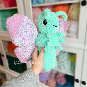 K&ouml;nnte beinhalten: Ein geh&auml;keltes Schmetterlings-Pl&uuml;schtier mit gr&uuml;nem K&ouml;rper und rosa-wei&szlig;en Fl&uuml;geln. Das Spielzeug hat ein s&uuml;&szlig;es Gesicht mit schwarzen Augen und einer rosa Nase. Das Spielzeug wird von einer Hand vor einem Garnregal gehalten.