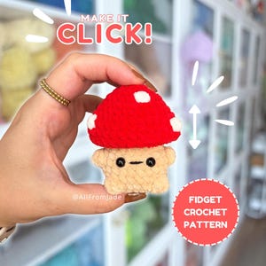 May include: A small, plush, crocheted mushroom toy with a bright red cap featuring white spots and a beige, square-shaped base with black bead eyes and a simple stitched mouth. The image includes the text "MAKE IT CLICK!" and "FIDGET CROCHET PATTERN".