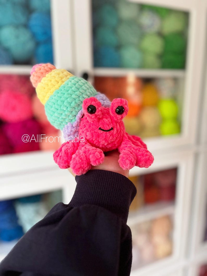 May include: A crocheted pink hermit crab toy with a rainbow shell. The crab has black eyes and a smiling mouth. The toy is held in a hand wearing a black sweater.