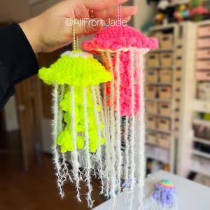 Peut inclure: Deux ornements de méduses faits à la main au crochet. L'un est vert fluo, l'autre rose vif. Les deux ont des franges blanches et sont suspendus par des chaînes dorées. Le texte "@AllFromJade" est visible.