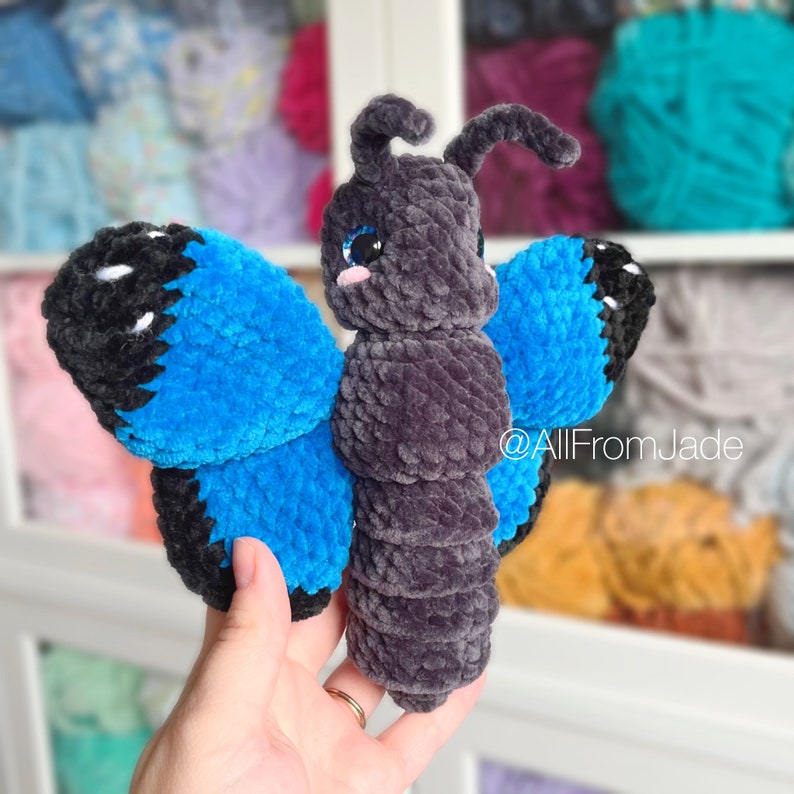 K&ouml;nnte beinhalten: Ein geh&auml;keltes Schmetterlings-Spielzeug mit blauen Fl&uuml;geln und einem grauen K&ouml;rper. Der Schmetterling hat schwarze Augen und eine rosa Nase. Das Spielzeug wird in einer Hand gehalten.