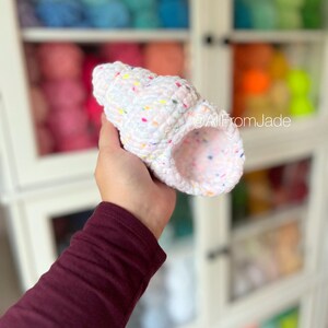 May include: White crocheted seashell with colorful speckles. The seashell is held in a hand.