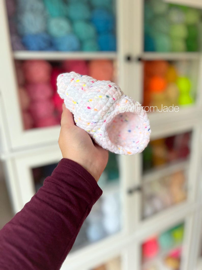 May include: White crocheted seashell with colorful speckles. The seashell is held in a hand.