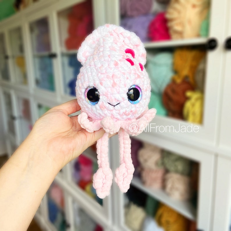 K&ouml;nnte beinhalten: Ein handgefertigtes, geh&auml;keltes Tintenfisch-Pl&uuml;schtier in Rosa und Wei&szlig; mit gro&szlig;en, glitzernden Augen und einem l&auml;chelnden Gesicht. Das Pl&uuml;schtier hat lange, herabh&auml;ngende Tentakel und ist mit kleinen rosa Akzenten verziert. Der Text "@AllFromJade" ist sichtbar.