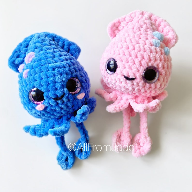 K&ouml;nnte beinhalten: Zwei handgefertigte H&auml;keltier-Tintenfische. Einer ist blau, der andere rosa. Beide haben gro&szlig;e, ausdrucksstarke Augen, kleine M&uuml;nder und Tentakel. Der blaue Tintenfisch hat lila Akzente, der rosa hellblaue. Auf wei&szlig;em Hintergrund.