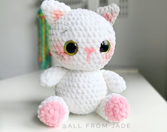 May include: A handmade white crochet cat plushie with pink accents on the ears, cheeks, and paws. The cat has large, expressive green eyes. The plushie is sitting on a white surface. The text "@ALL FROM JADE" is at the bottom.