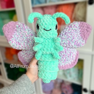 K&ouml;nnte beinhalten: Ein geh&auml;keltes Schmetterlings-Pl&uuml;schtier mit gr&uuml;nen und rosa Fl&uuml;geln. Der Schmetterling hat einen gr&uuml;nen K&ouml;rper mit schwarzen Augen und einem l&auml;chelnden Mund. Die Fl&uuml;gel sind aus einem strukturierten Garn mit einem wei&szlig;en und rosa Muster gefertigt. Das Spielzeug wird in einer Hand gehalten, wobei der Instagram-Handle @AllFromJade in der linken unteren Ecke sichtbar ist.